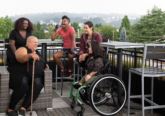 A multiracial group of women talking and laughing around a table on a patio. One is a wheelchair user, another uses a cane. 
